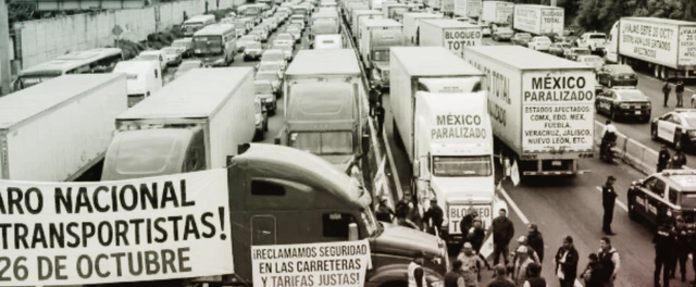 Fila de tractocamiones detenidos en una autopista con una caseta al fondo y conductores junto a sus unidades